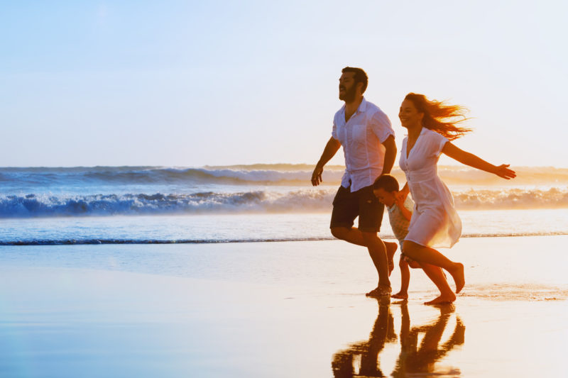 Happy family – father, mother, baby son hold hands and run together with fun along sunset sea surf on black sand beach. Travel, active lifestyle, parents with children on tropical summer vacations. happy family